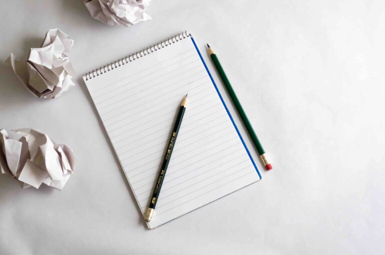 Spiral notepad with pencils and crumpled paper balls scattered on a white desk.