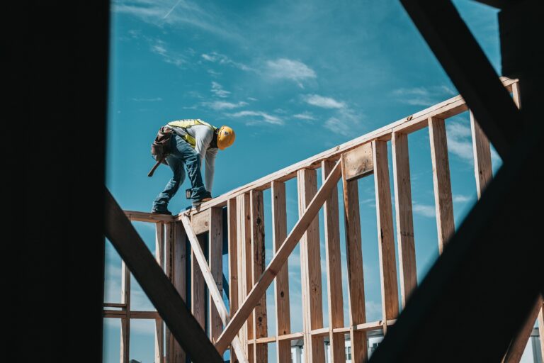 Construction worker in safety gear bending over wooden roof framing under blue sky.