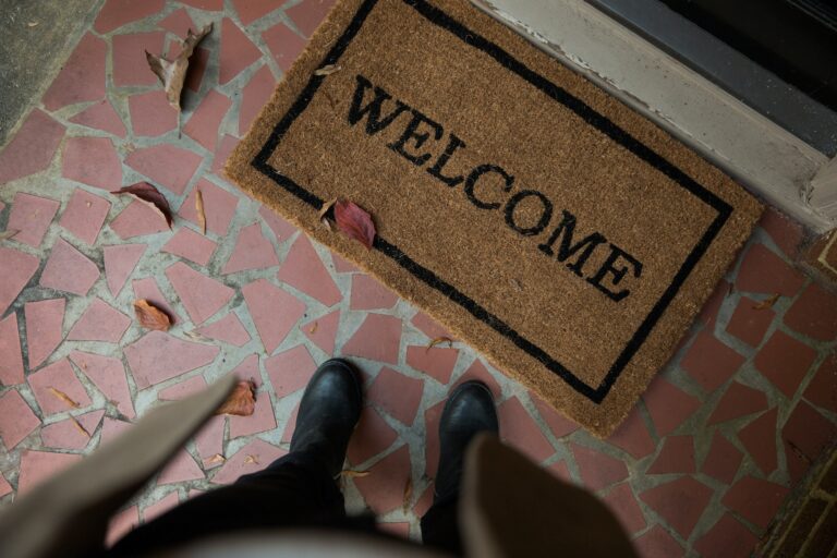 Black boots standing on a Welcome doormat over tiled porch with scattered autumn leaves.
